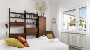 Een slaapkamer met een bed, een houten boekenkast, een radiator en een raam met uitzicht op gebouwen.