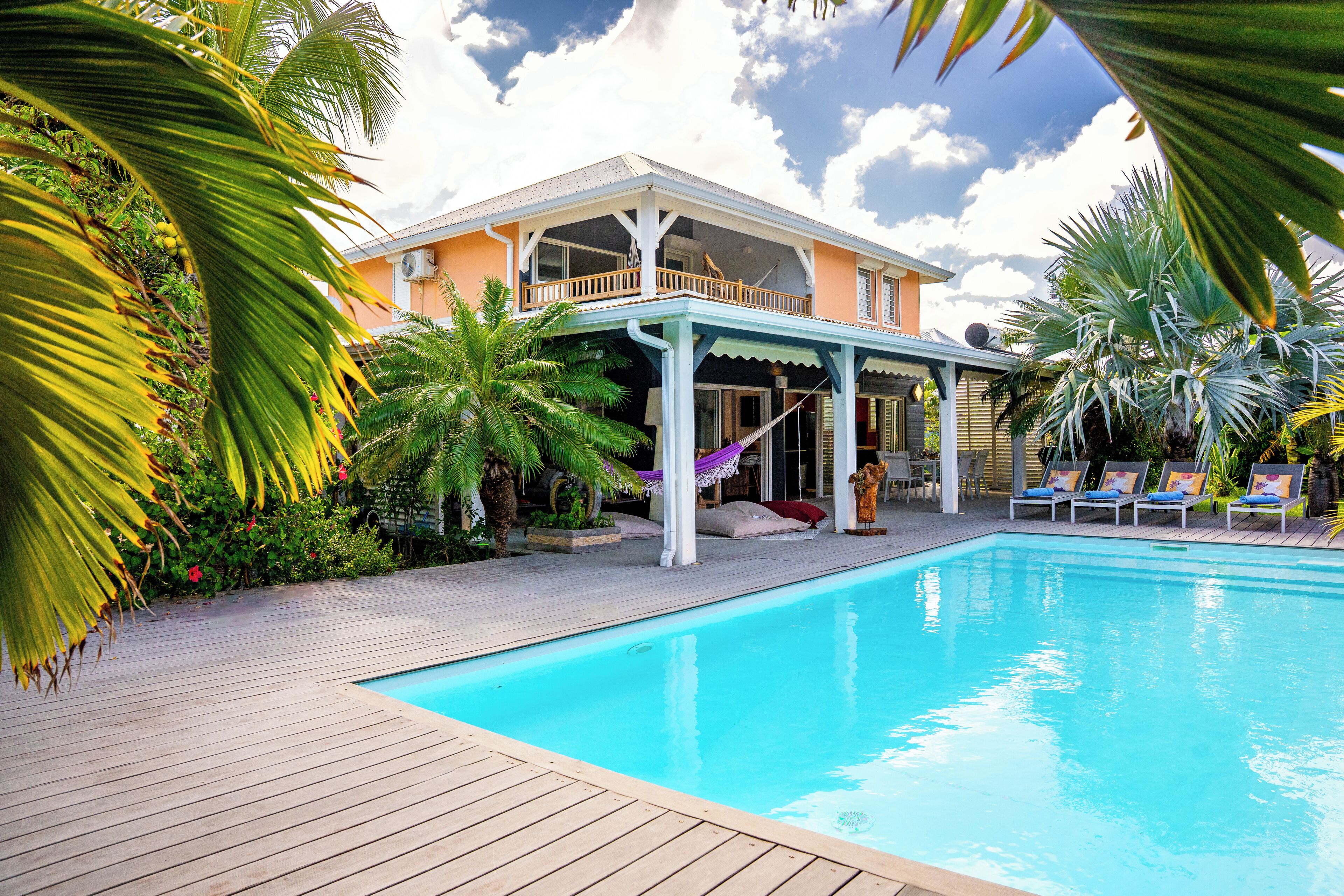 Un espace aménagé au bord de la piscine, avec une terrasse en bois, des chaises longues et un patio couvert offrant une vue sur une maison.