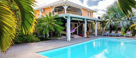 Un espace aménagé au bord de la piscine, avec une terrasse en bois, des chaises longues et un patio couvert offrant une vue sur une maison.