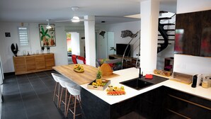 A modern kitchen with a central island, a dining area with chairs, a staircase, and a large TV.