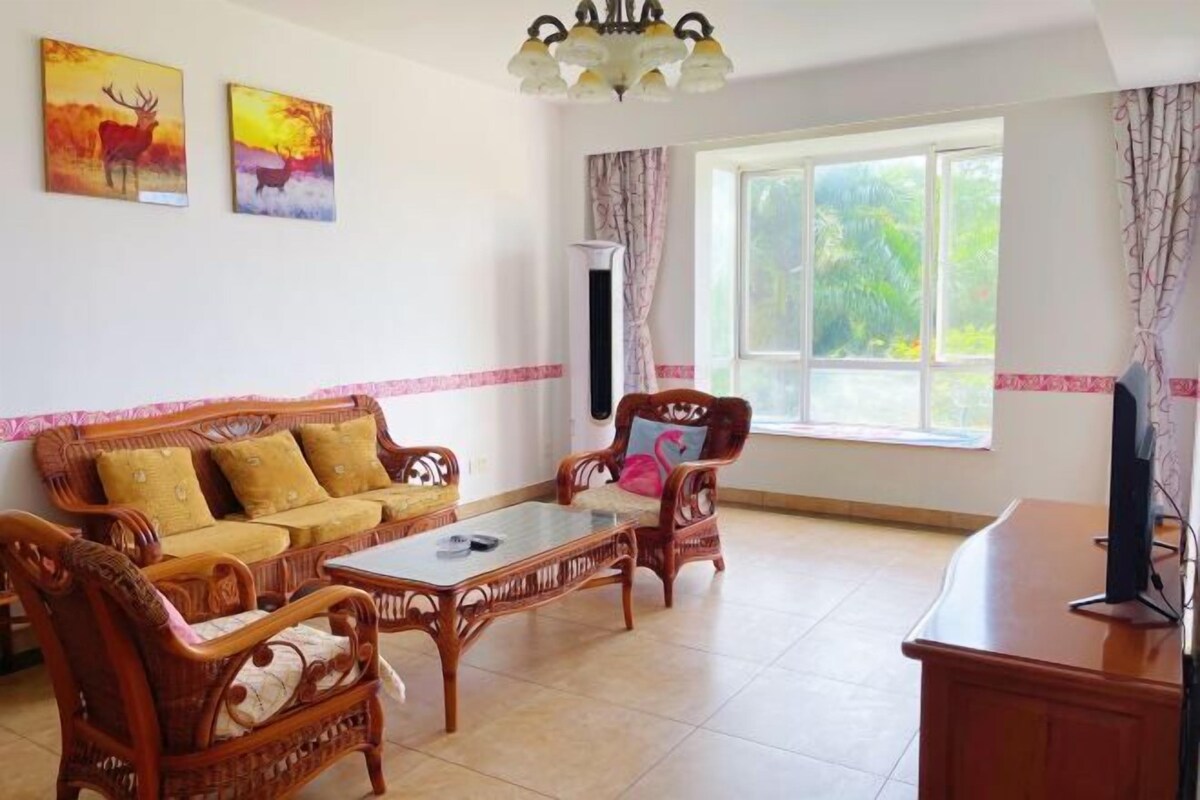 Un salon comprenant un canapé, des fauteuils, une table basse et un téléviseur. Deux tableaux ornent le mur, et une fenêtre est agrémentée de rideaux.