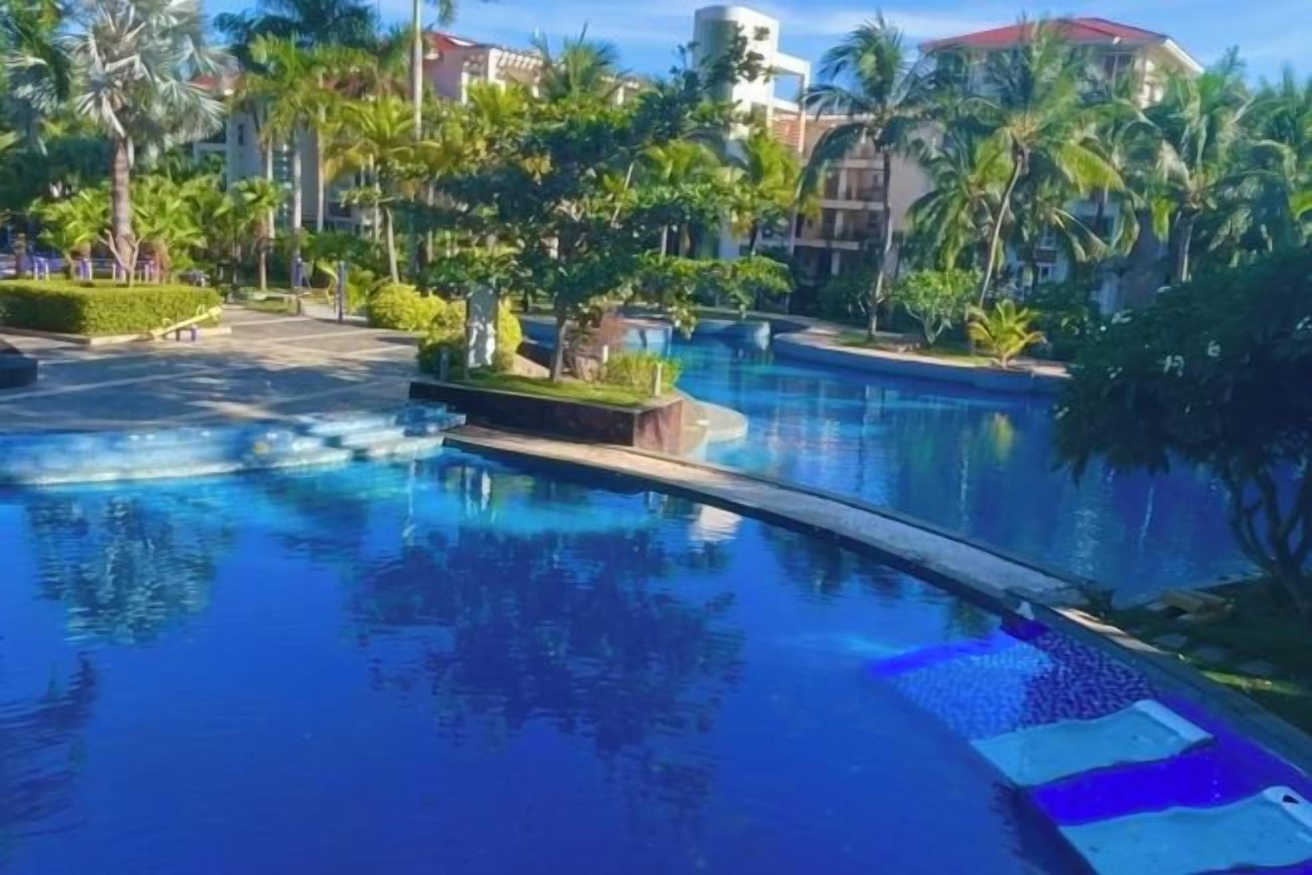 Una piscina de borde curvo, rodeada de palmeras y vegetación, con edificios al fondo.