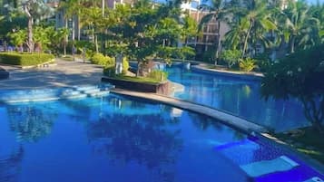 A swimming pool with a curved edge, surrounded by palm trees and greenery, with buildings in the background.