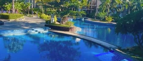 A swimming pool with a curved edge, surrounded by palm trees and greenery, with buildings in the background.