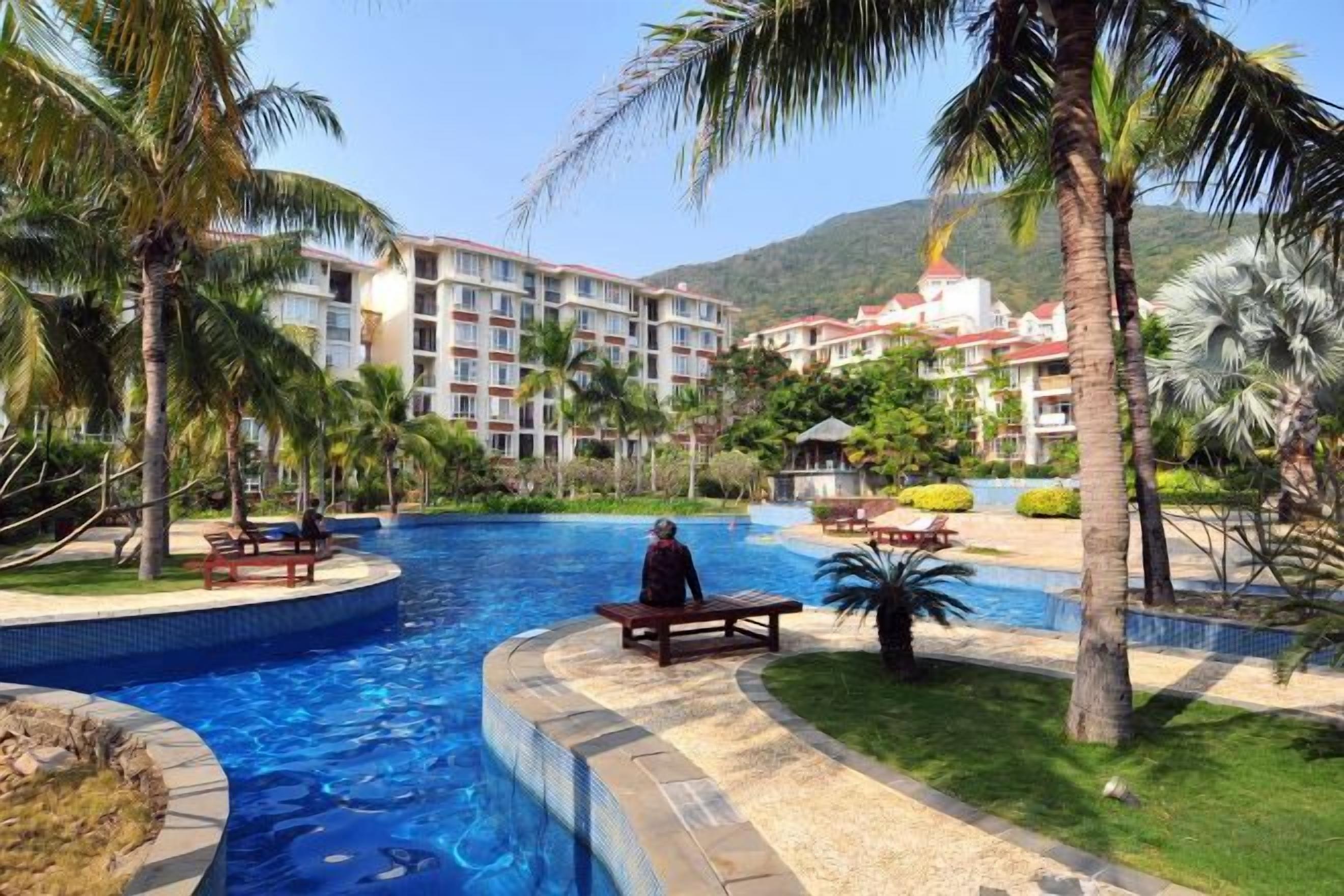 Una piscina de borde curvo, rodeada de palmeras y un edificio con varios balcones al fondo.