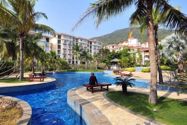 Una piscina de borde curvo, rodeada de palmeras y un edificio con varios balcones al fondo.