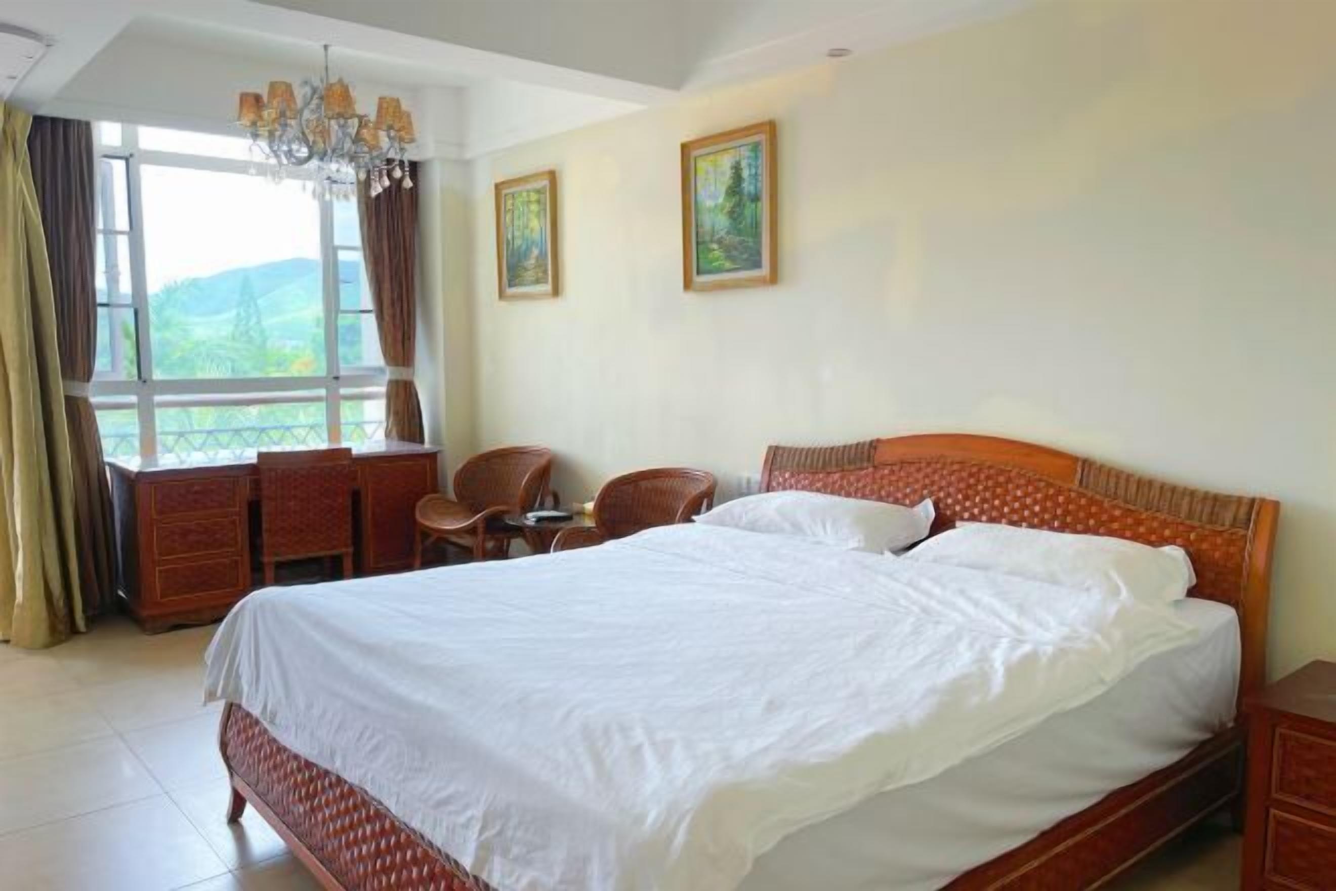 A bedroom with a large bed, a desk, and two chairs. There are two paintings on the wall and a window with curtains.