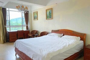 Une chambre avec un grand lit, un bureau et deux chaises. Il y a deux tableaux au mur et une fenêtre avec des rideaux.
