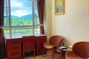 Une pièce avec vue sur des collines verdoyantes, un bureau en bois, deux fauteuils en osier et une petite table sur laquelle se trouvent un livre et une boîte.