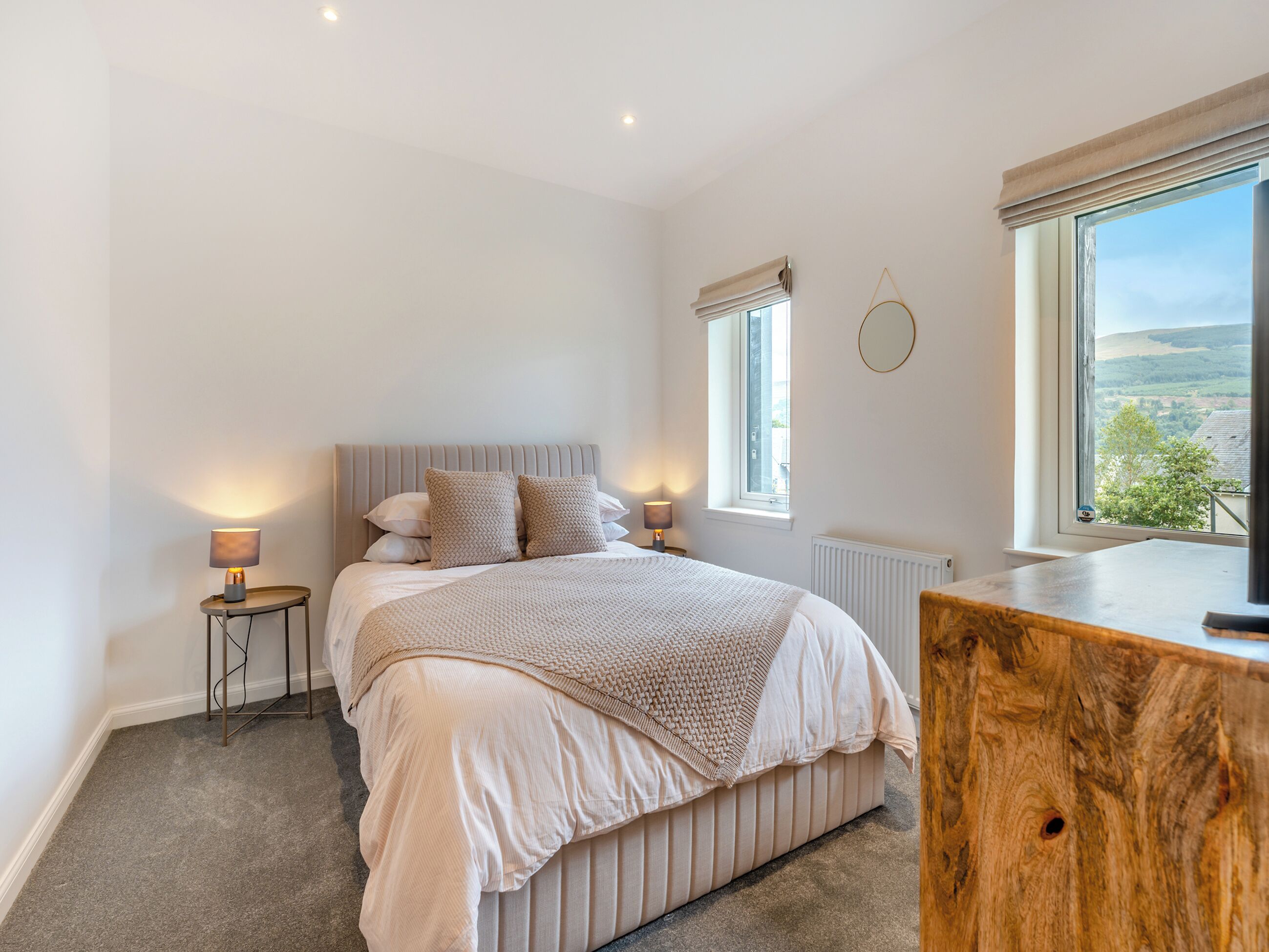 A bedroom with a bed, bedside tables, a wooden dresser, a round mirror, and a window with a view.