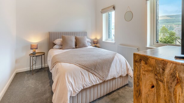 Une chambre à coucher comprenant un lit, des tables de chevet, une commode en bois, un miroir rond et une fenêtre donnant sur un paysage.