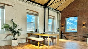Une pièce spacieuse avec un plancher en bois, une grande table entourée de chaises, une plante en pot et une grande fenêtre donnant sur un immeuble.