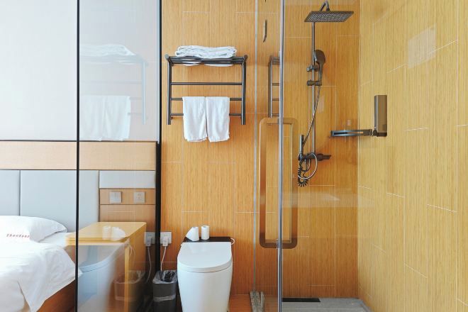 A modern bathroom with a glass shower enclosure, a wooden vanity, and a bed with a white headboard.