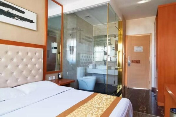 A hotel room with a bed, a glass-enclosed shower, a bathroom with a mirror, and a door.