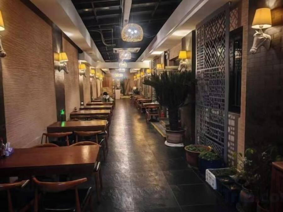 A restaurant with a long counter, wooden tables, and chairs, and wall-mounted lamps.