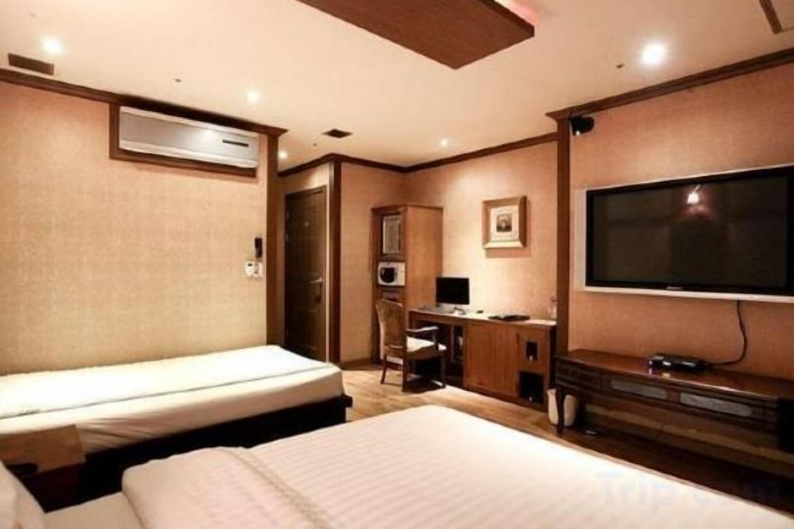 A hotel room with two beds, a desk, a television, and an air conditioning unit.