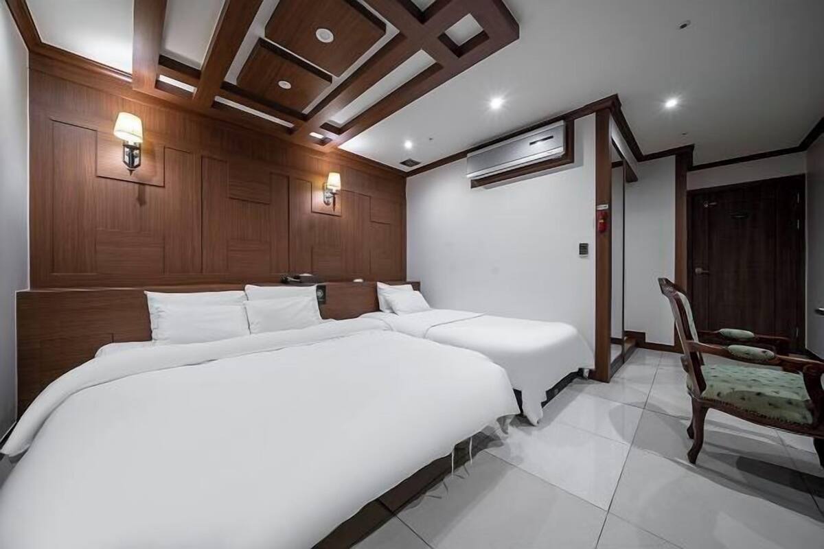 Une chambre d’hôtel avec deux lits, une tête de lit en bois, des lampes fixées au mur et un appareil de climatisation.