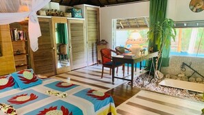 A room with a bed, a table, a chair, a small table with a plant, and a window with a view of the outdoors. - Polynesian Bungalow"seashell" Private Bath AC with shared swimming pool (Moorea-Maiao)