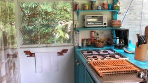 A compact kitchen with a gas stove, a sink, and a window with a view of greenery. There are shelves with jars and a toaster oven. - Polynesian Bungalow"seashell" Private Bath AC with shared swimming pool (Moorea-Maiao)