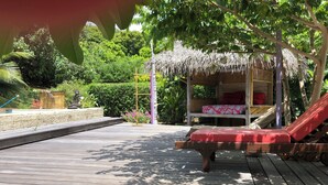 A wooden deck with a thatched-roof structure, a red cushioned lounge chair, and a statue. - Polynesian Bungalow"seashell" Private Bath AC with shared swimming pool (Moorea-Maiao)