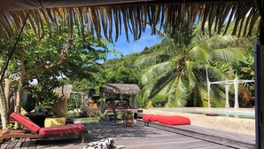A wooden deck with red and white patterned cushions, a thatched-roof structure, and a pool area with a white bridge. - Polynesian Bungalow"seashell" Private Bath AC with shared swimming pool (Moorea-Maiao)