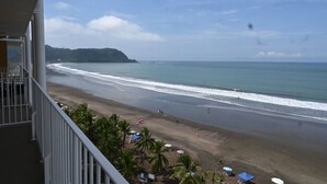 A beach view from a balcony with a clear sky, palm trees, and a distant hill. - Jacó beachfront condo in center of town (Jaco)