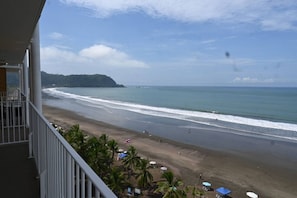 A beach view from a balcony with a clear sky, palm trees, and a distant hill.