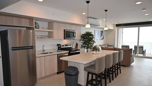 A modern kitchen with a stainless steel refrigerator, microwave, and stove. There is a dining area with a bar stool and a large window. - Jacó beachfront condo in center of town (Jaco)