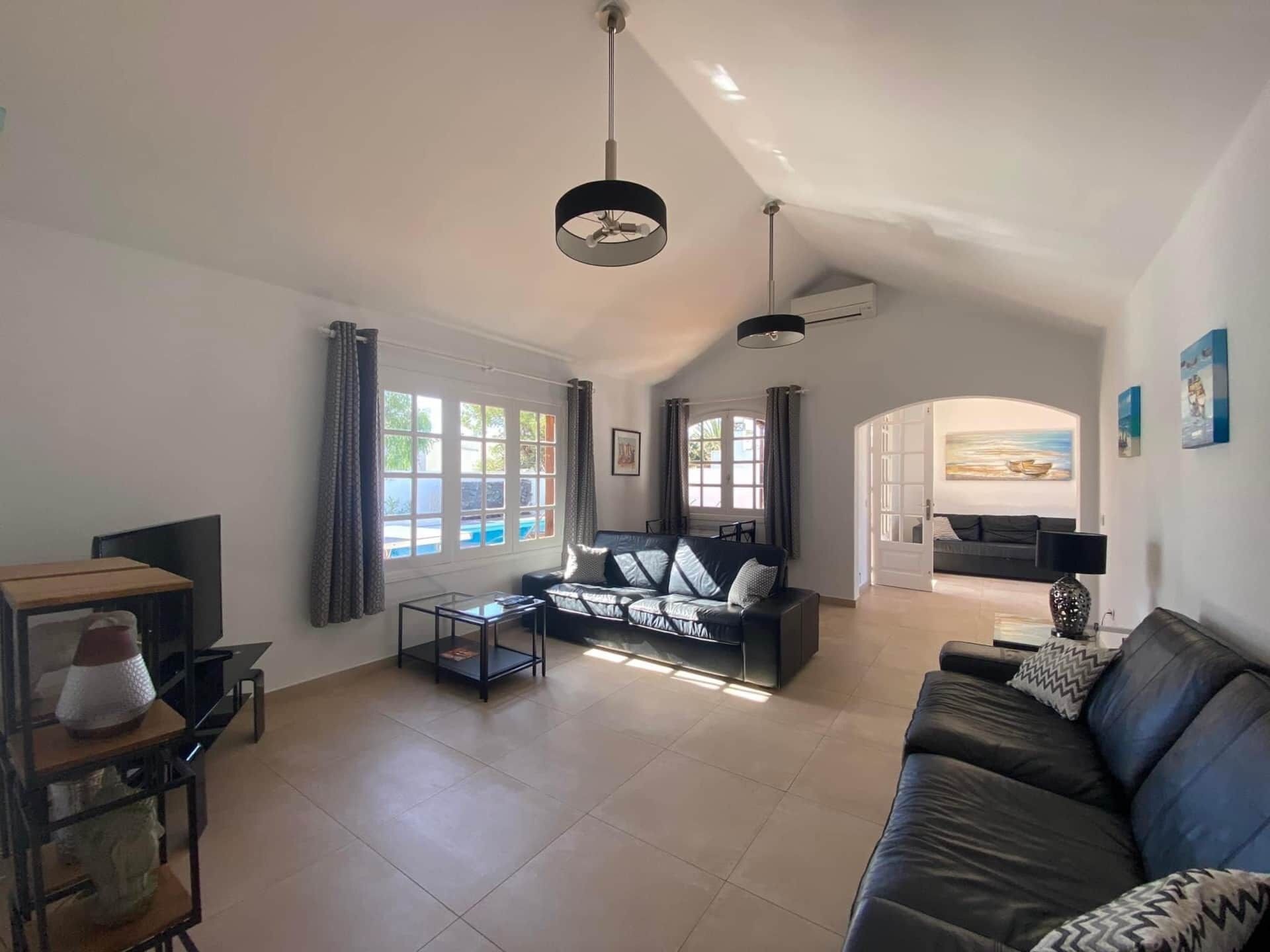 A spacious living room with a large black leather sofa, a glass coffee table, and a TV on a wooden stand. There are two windows with curtains, a ceiling fan, and a painting on the wall.