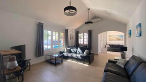 A spacious living room with a large black leather sofa, a glass coffee table, and a TV on a wooden stand. There are two windows with curtains, a ceiling fan, and a painting on the wall. - Villa Alicia, Moderne Exklusive Villa in Puerto del Carmen (Tías)