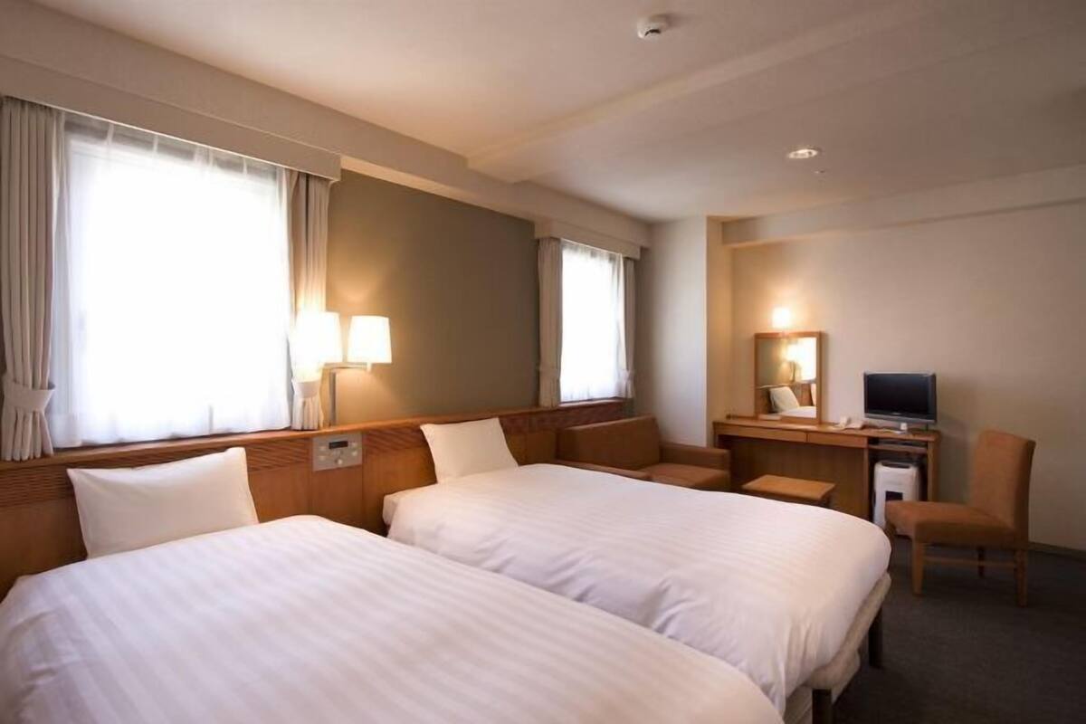 A hotel room with two beds, a desk with a TV, a chair, and a large window with curtains.