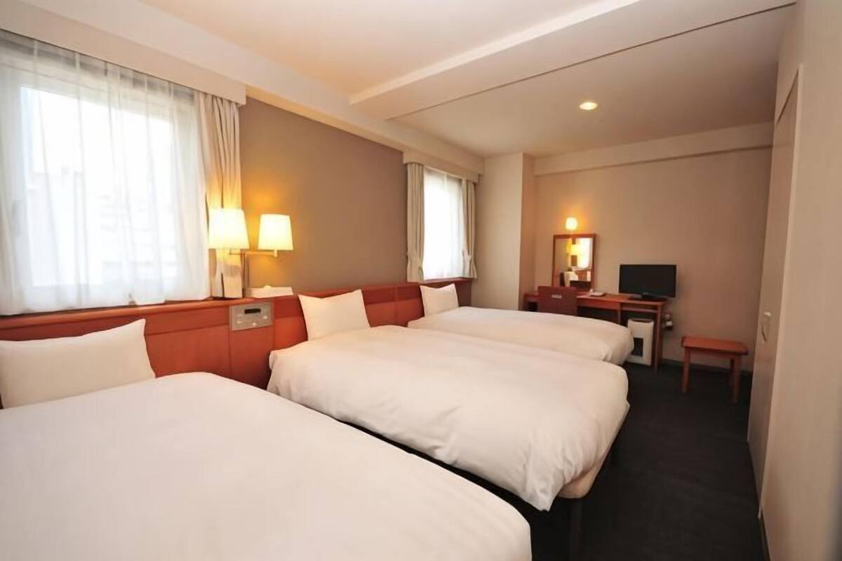 A hotel room with three beds, a desk, a television, and a window with curtains.