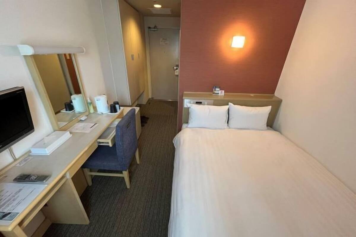 A hotel room with a desk, chair, bed, and a wall-mounted light.
