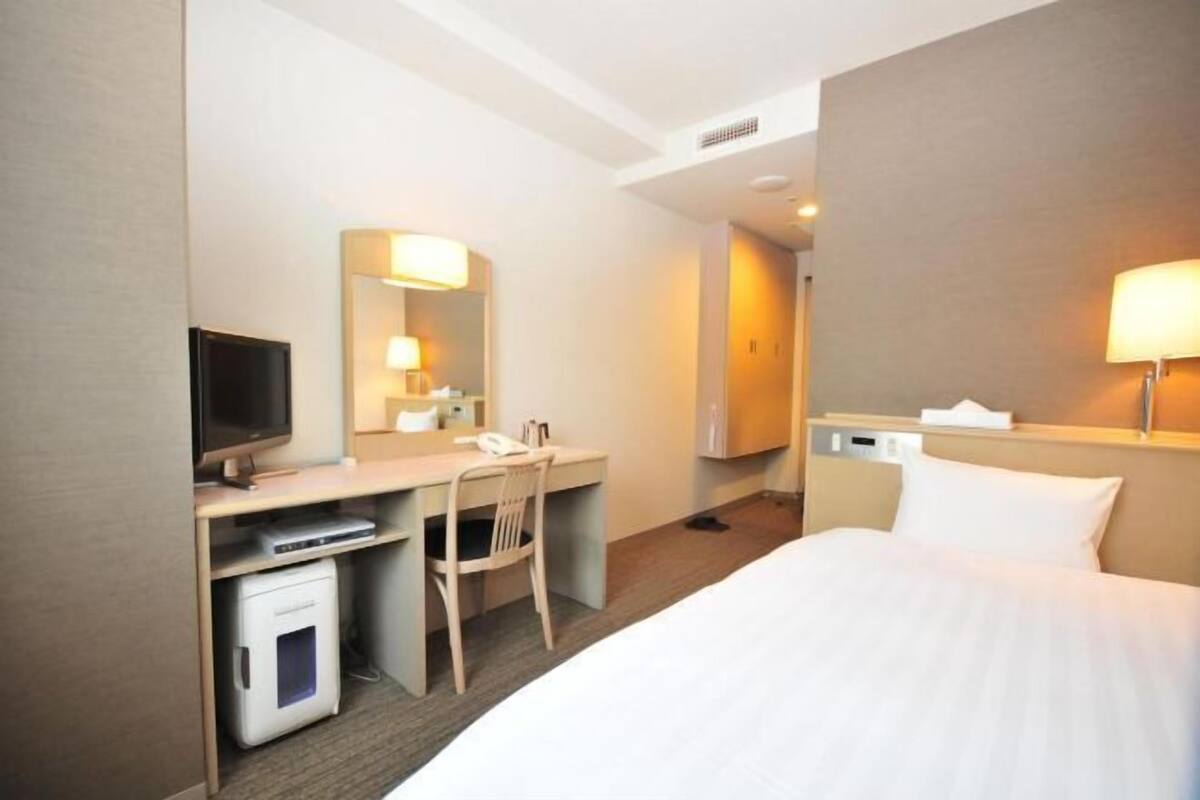 A hotel room with a bed, a desk with a chair, a television, a mirror, and a bedside table with a lamp.