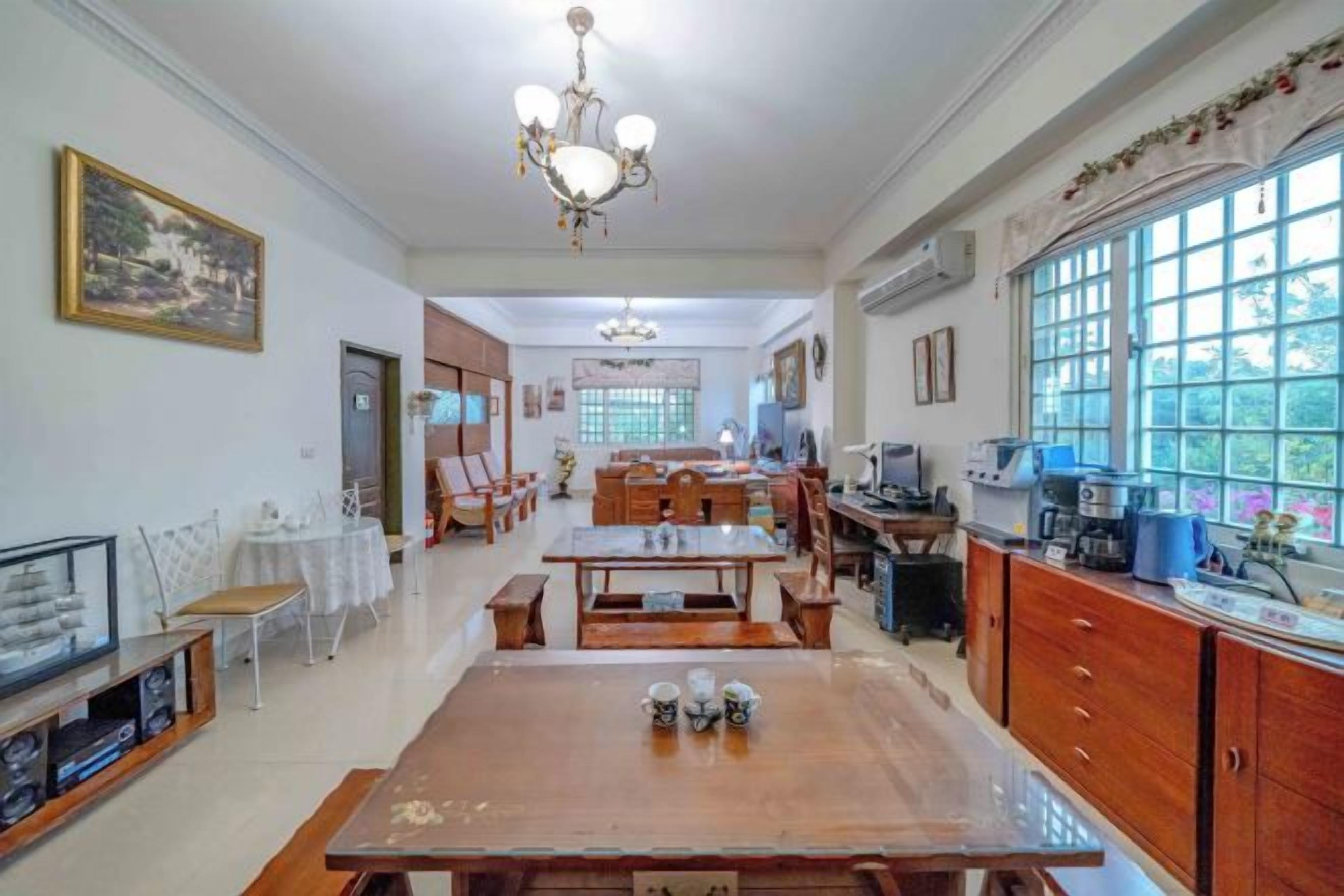 Un'ampia sala da pranzo con un grande tavolo in legno, sedie e un lampadario. È presente anche un angolo cottura con pensili e una finestra.