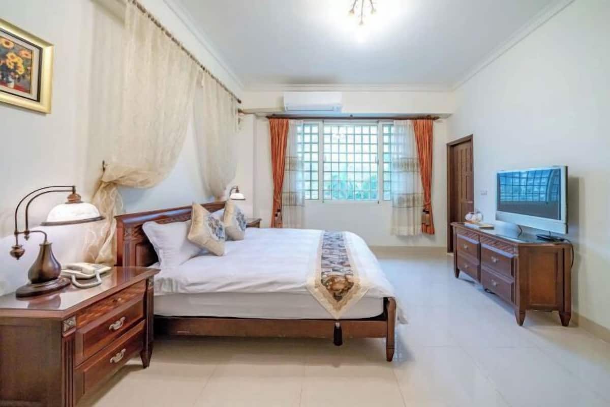 A bedroom with a bed, bedside tables, an air conditioning unit, a television, and curtains.