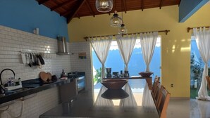 A modern kitchen with a black countertop, white tiled backsplash, and a large island. There are hanging lights, a view of the outdoors, and a dining area with a table and chairs. - Big house with 4 bedrooms, 4 1/2 bath, barbecue area,. Pool, Jacuzzi and parking (Arraial d'Ajuda)