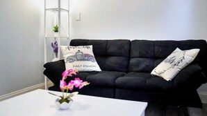 Een zwarte bank met twee decoratieve kussens, waarvan één een vrachtwagenprint heeft en de tekst 'farm life DAILY Deliveries', en een witte bijzettafel met een vaas vol roze bloemen.