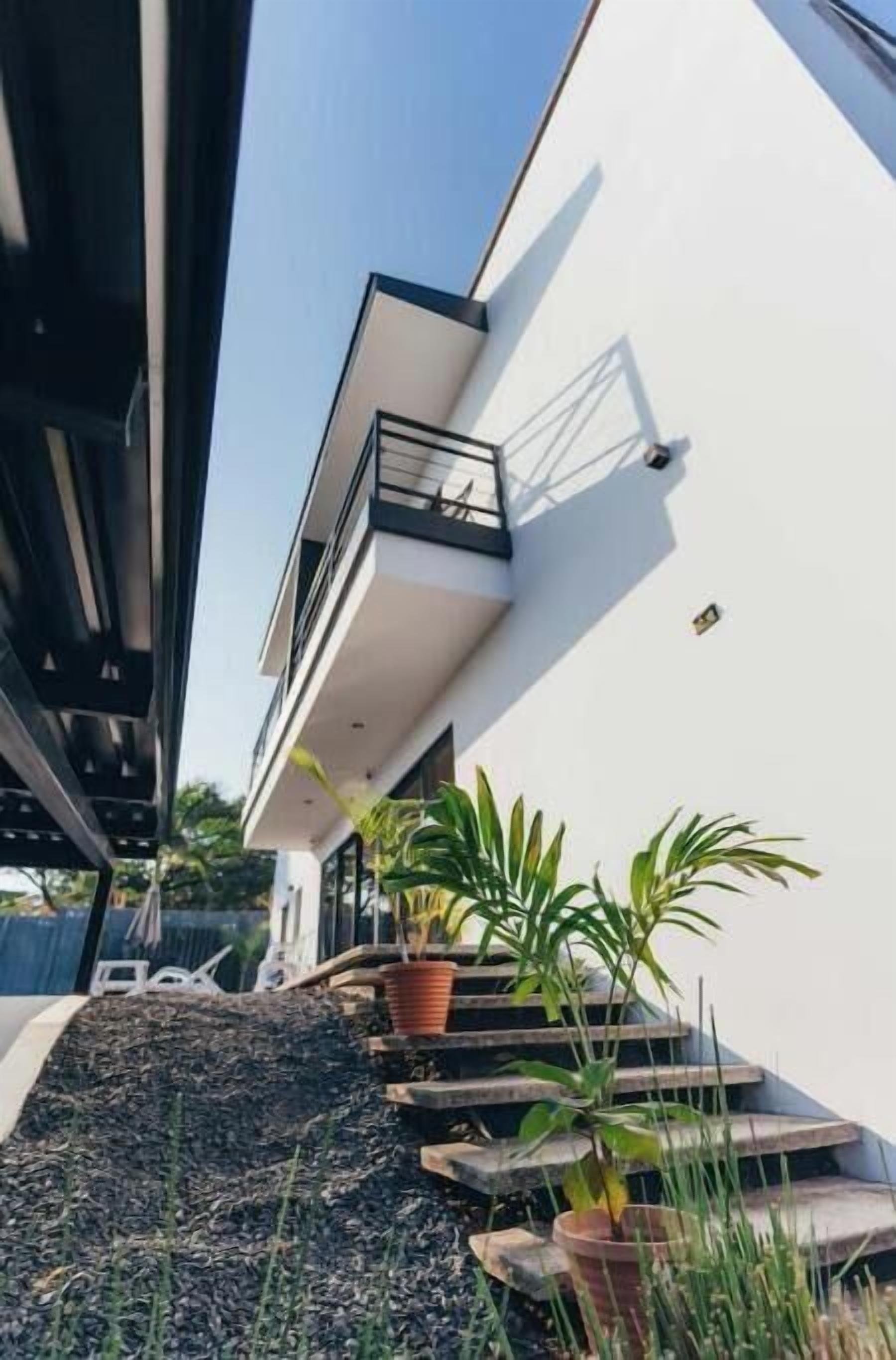 Il s’agit d’un bâtiment moderne à deux étages, doté d’une façade blanche et d’un balcon. On y trouve des plantes en pot et un petit escalier menant à l’entrée.