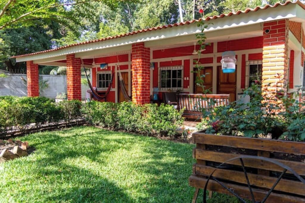 A red brick building with a porch, a small garden, and a wooden bench.