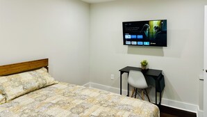 A bedroom with a bed, a desk, a chair, a ceiling fan, and a mounted TV displaying a show.