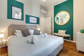 A neatly made bed with pillows and towels, a bedside table with a lamp, a round mirror, and a small bench. - Mamo Florence - Bice & Krizia Apartments (Florence)