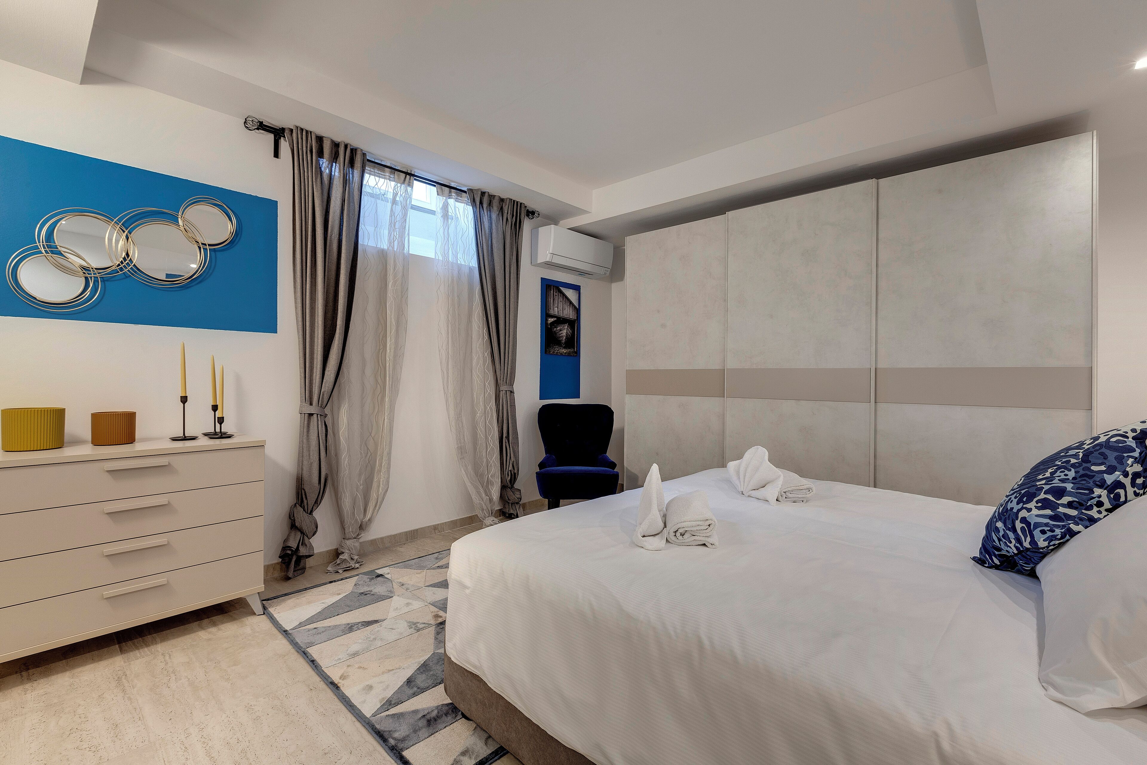 Moderni hotellihuone, jossa on suuri sänky, lipasto, meikkipöytä kynttilöineen ja sininen tehosteseinä.