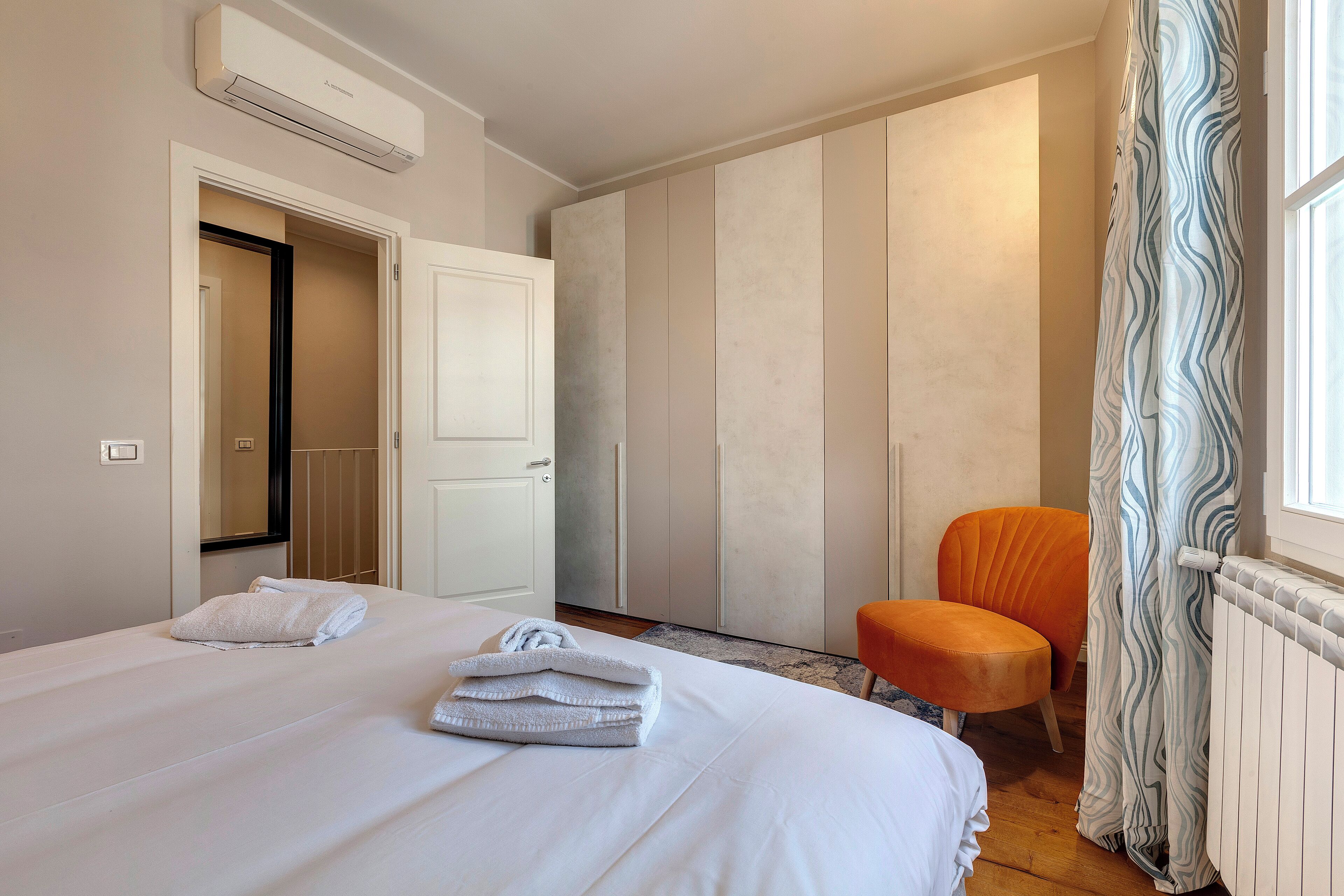 Una habitación de hotel con cama, una silla, armario y ventana con cortinas.
