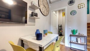 Ein Essbereich mit Tisch, Stühlen und einem an der Wand befestigten Fernseher. Regale mit Dekorationselementen und eine Tür zu einem weiteren Raum.