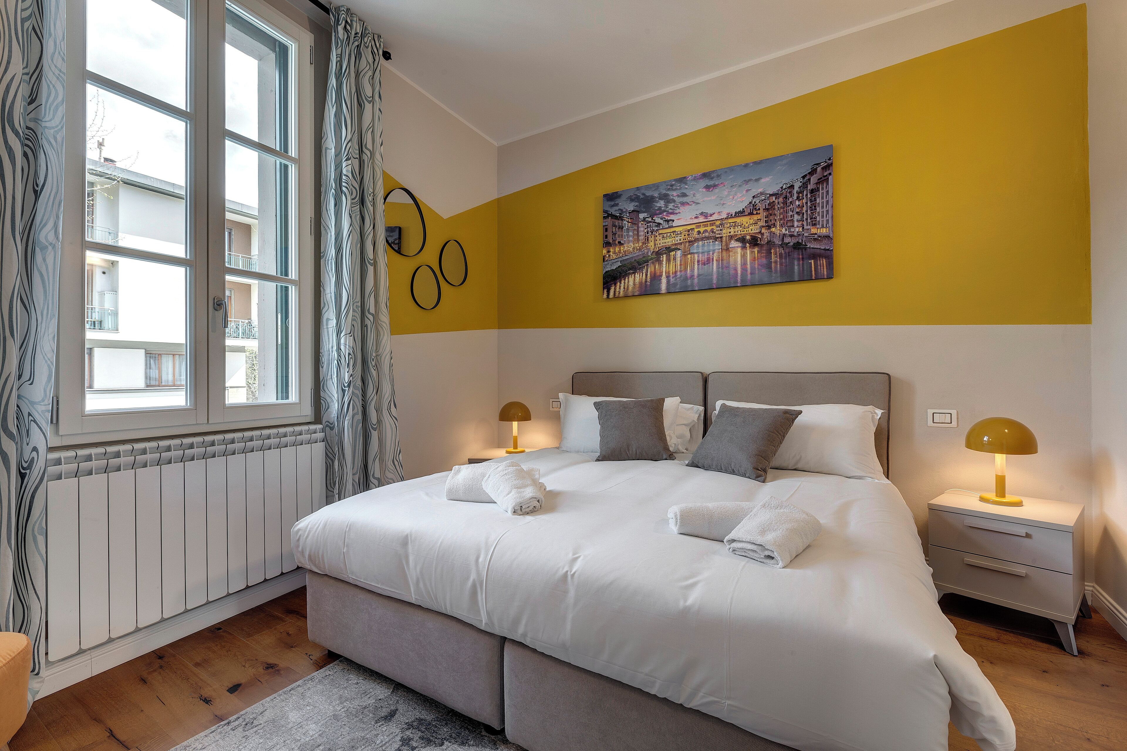 A neatly made bed with white linens, a bedside table with a lamp, and a cityscape painting on the wall.