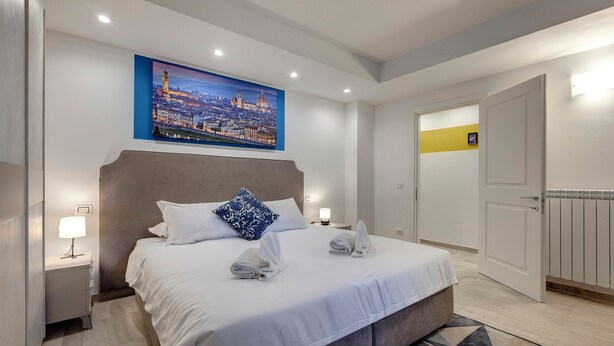 Ein Hotelzimmer mit einem großen Bett, einem Nachttisch mit Lampe, einem Stadtbild als Gemälde und einer Tür mit gelber Streifen.