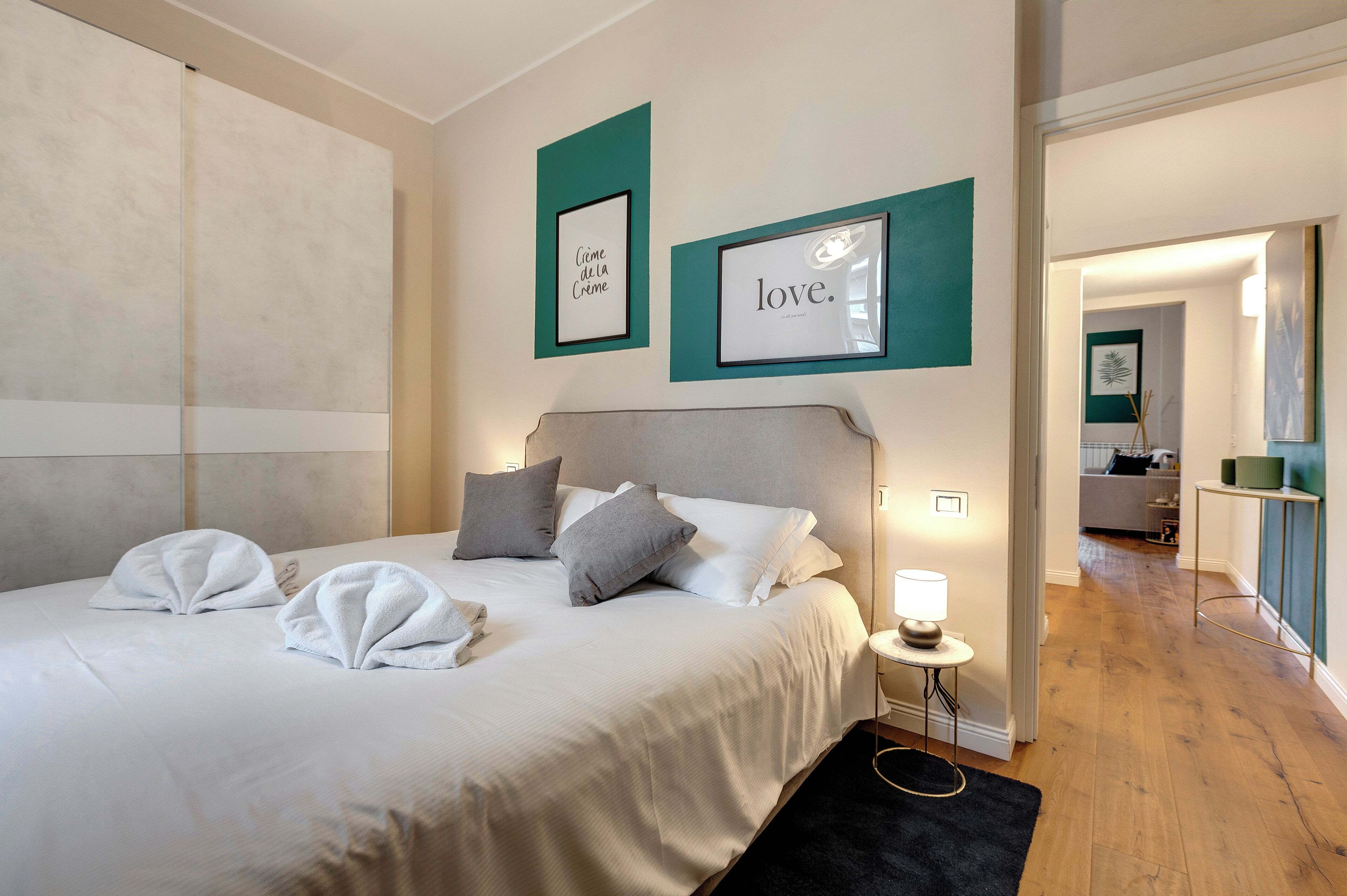 A neatly made bed with white linens and towels, a bedside table with a lamp, and two framed pictures on the wall.