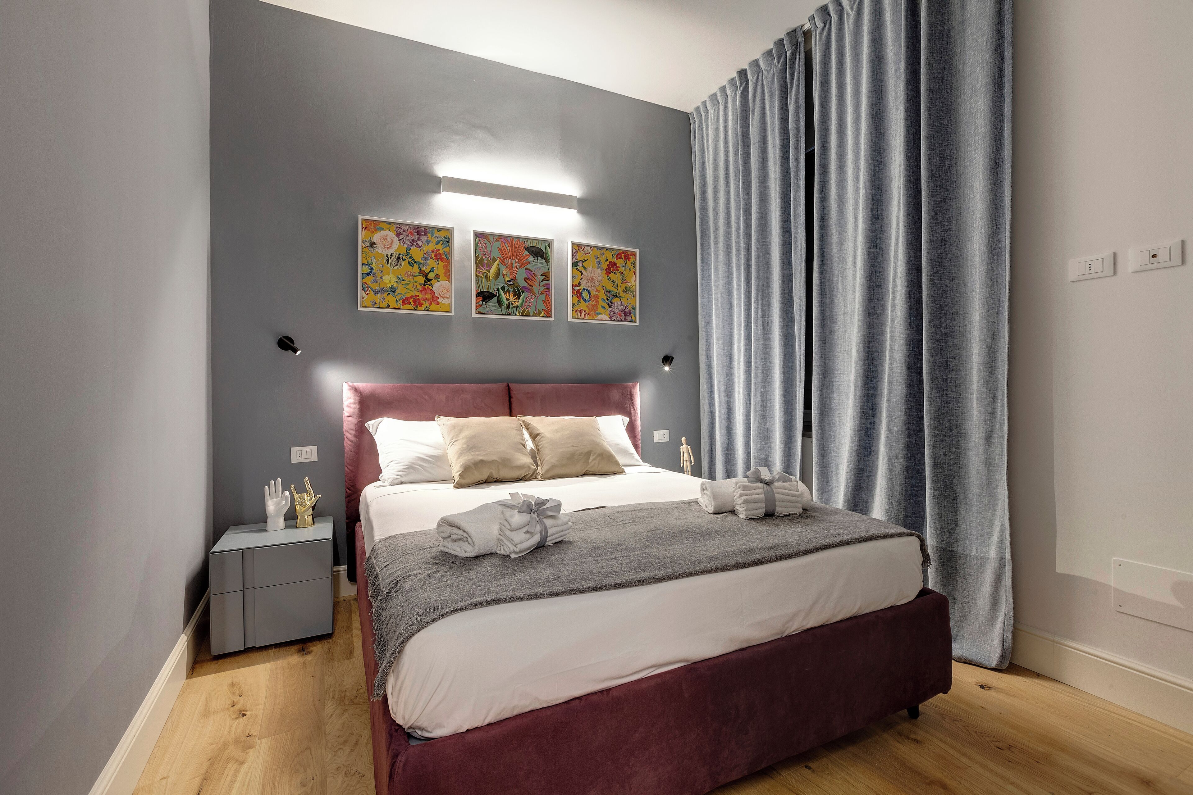 Ett modernt sovrum med en grå vägg, en röd sänggavel och ett grått sängöverkast. Ovanför sängen hänger tre tavlor med blommotiv, och bredvid sängen står ett nattduksbord med en lampa. Ett fönster med grå gardiner finns också i rummet.