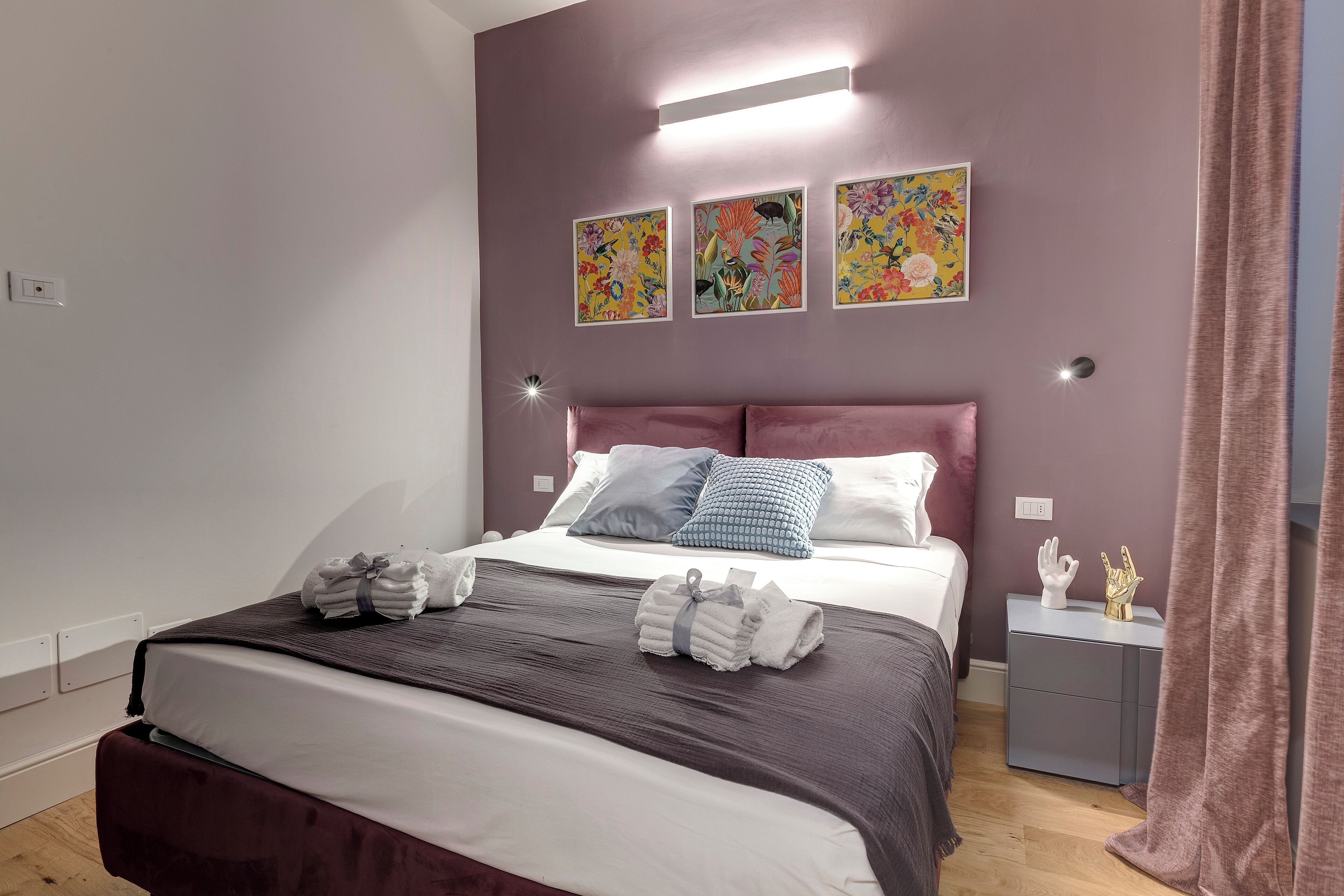 Una cama bien tendida con cabecera marrón, almohadas blancas y azules, y un juego de toallas. Hay tres obras de arte colgadas en la pared sobre la cama, y una mesita de noche con un objeto decorativo.